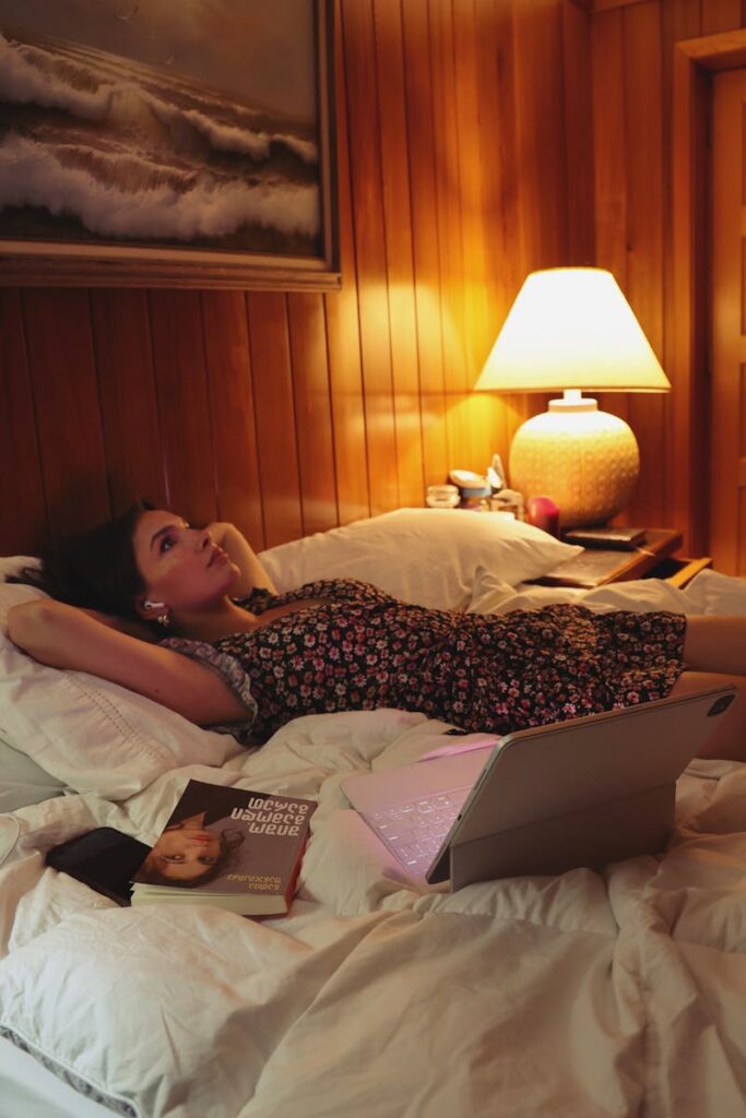 A woman in a floral dress lying on a bed with a book and a laptop nearby representing someone who is overthinking and ruminating for blog post.