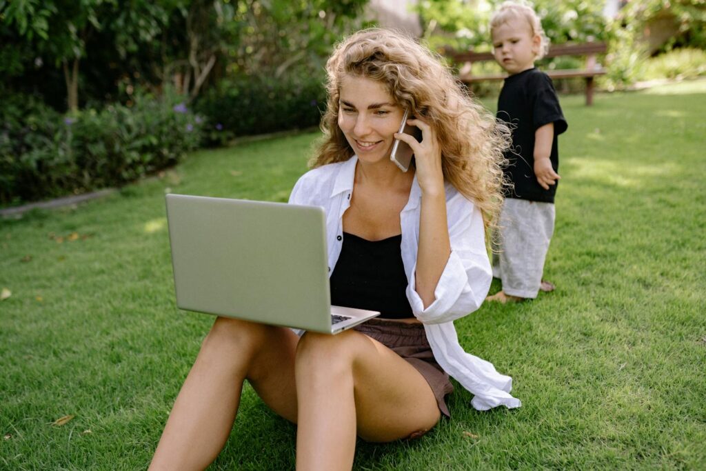 A woman multitasking with a laptop and phone while watching her child play in a sunny yard.